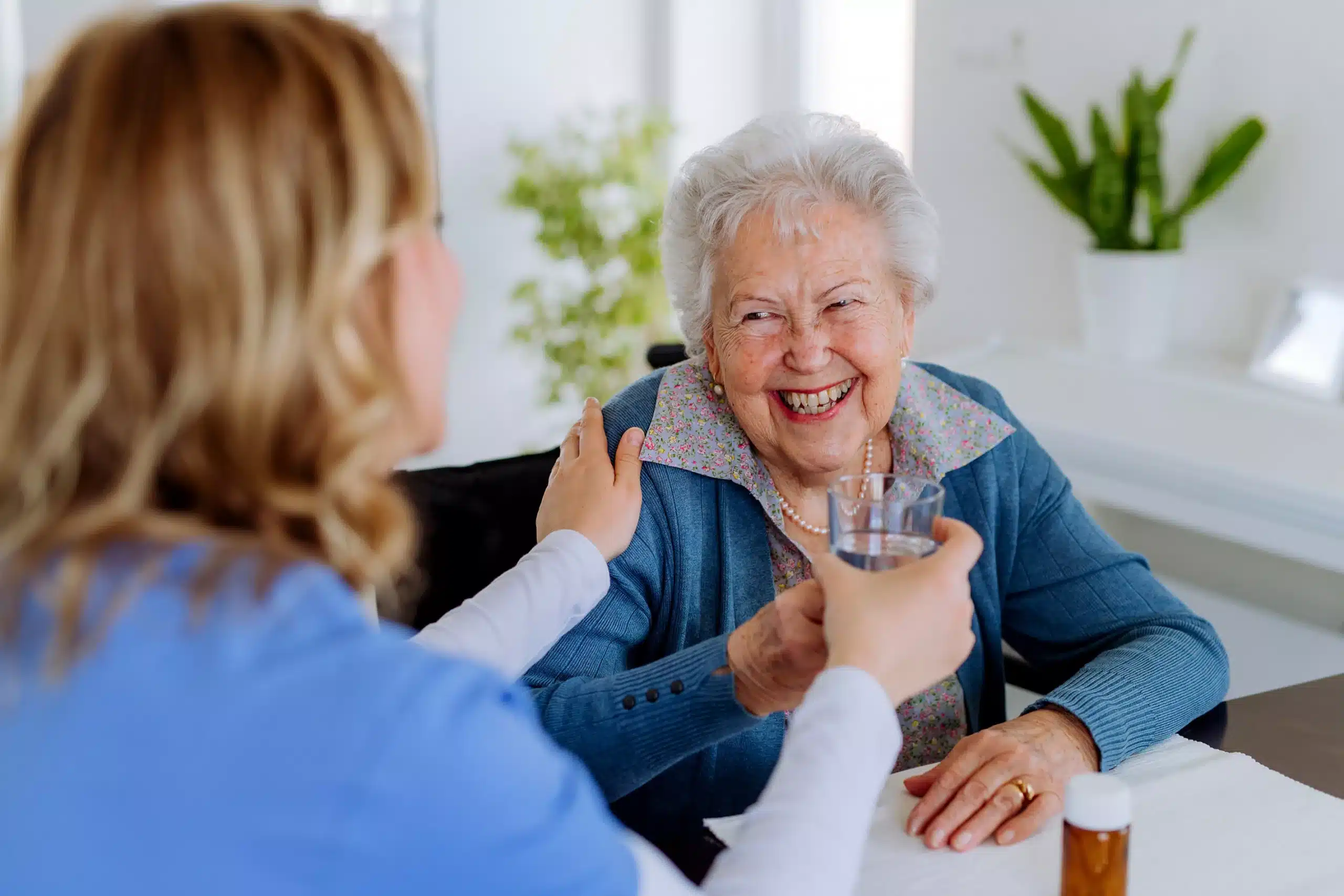 Ältere Dame wird von einer Pflegekraft untersucht und freundlich betreut. Pflegekraft reicht der Seniorin ein Wasserglas.