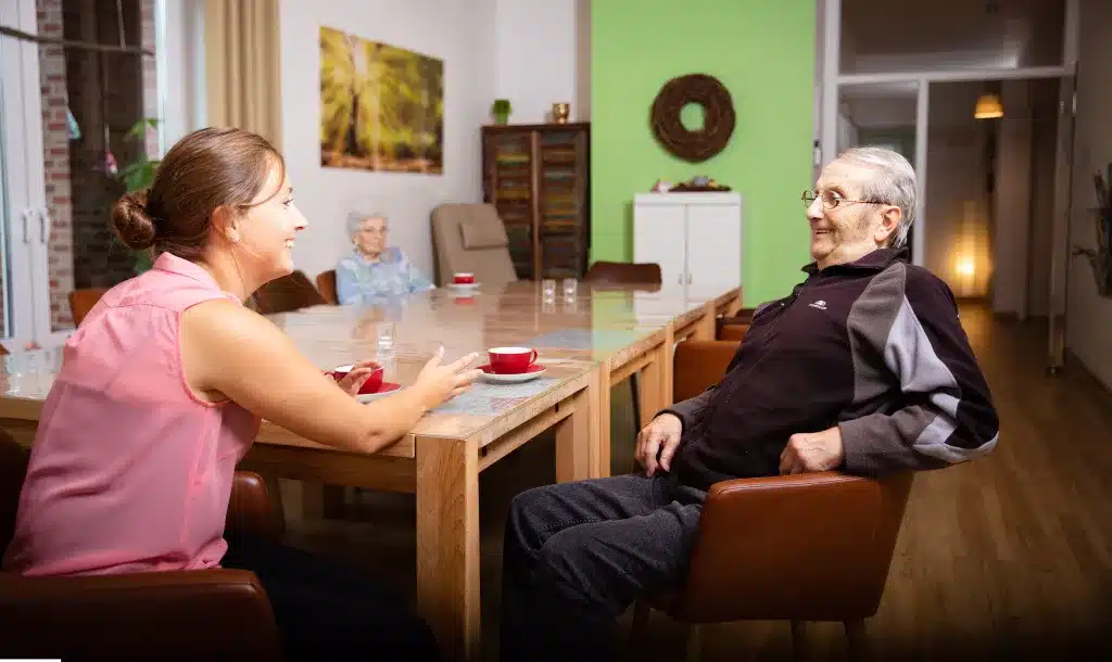 Eine Pflegekraft spricht mit einem Senioren am Tisch in der Tagespflegeeinrichtung der Firma Leusbrock Pflege.