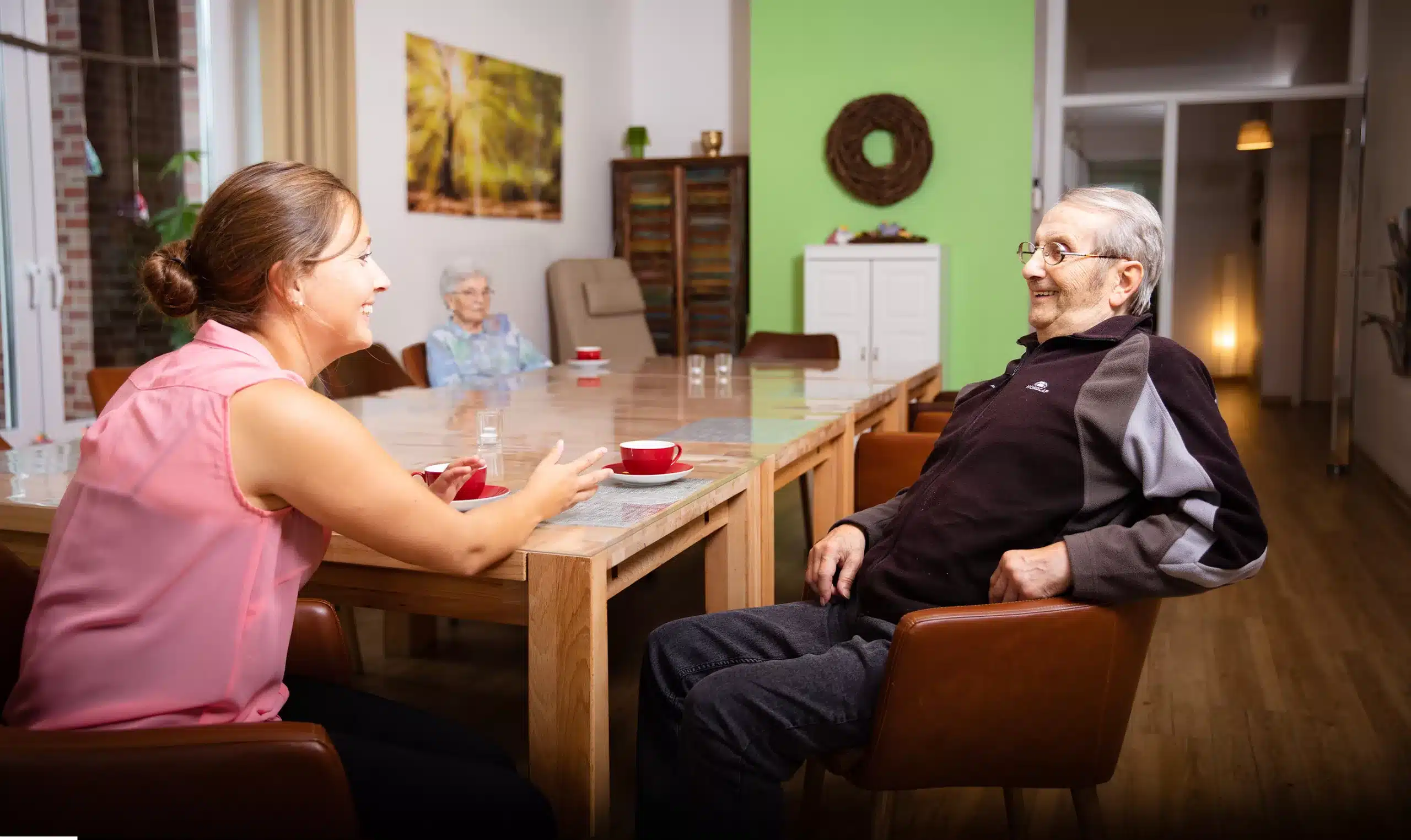 Eine Pflegekraft spricht mit einem Senioren am Tisch in der Tagespflegeeinrichtung der Firma Leusbrock Pflege.